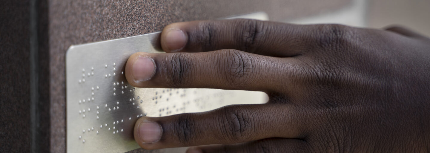 dedos deslizam sobre uma placa em braille afixada numa parede de uma rua