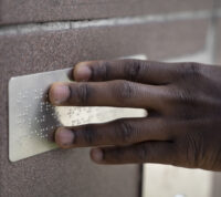 dedos deslizam sobre uma placa em braille afixada numa parede de uma rua