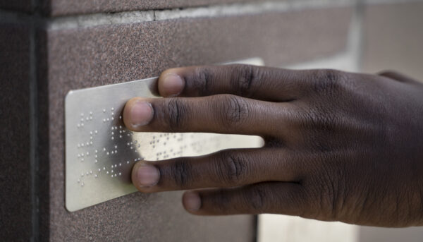 dedos deslizam sobre uma placa em braille afixada numa parede de uma rua